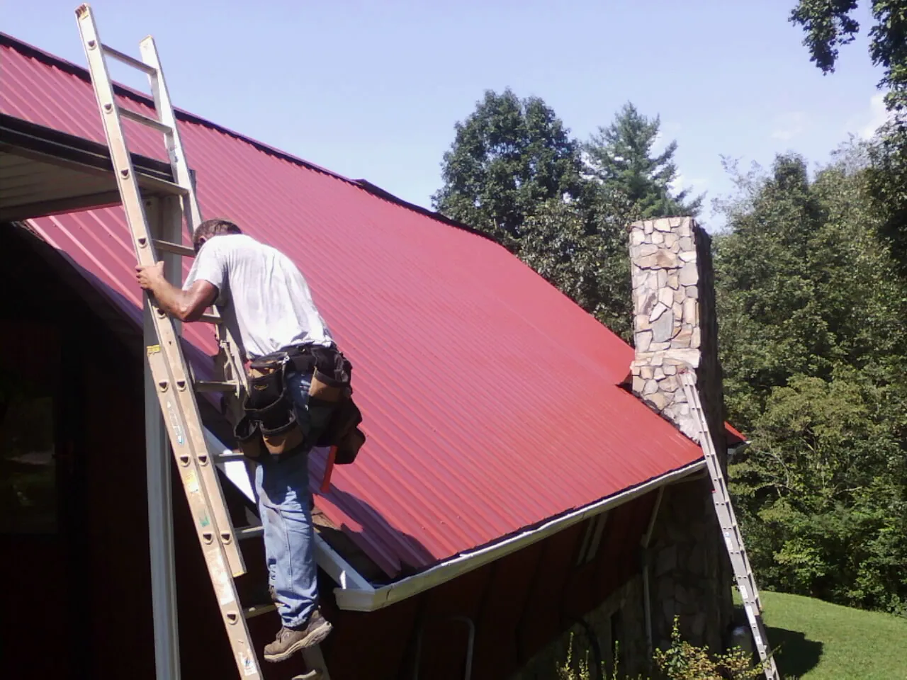 Roofer on a ladder beside a red metal roof and stone chimney — Soffit & Fascia Repair by Roofing Brothers in Dutch Flat